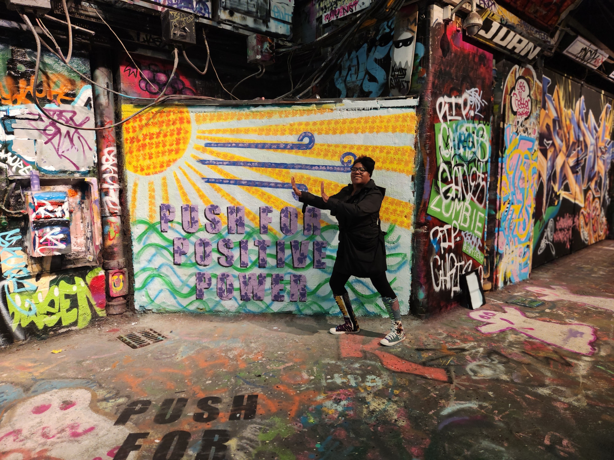 Dori LeGrand-Christmas standing beside a colorful Push for Positive Power mural in a graffiti-covered alley representing hope, strength, and forward movement